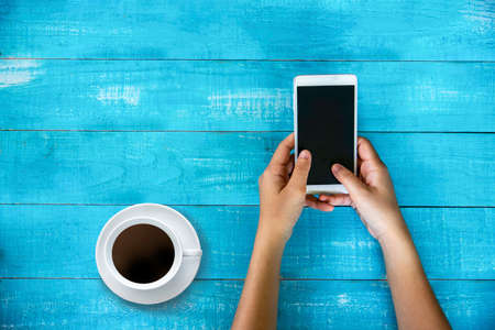 Hand holding mobile phone with blank screen with coffee cup over wood table. technology and lifestyle concept.の写真素材