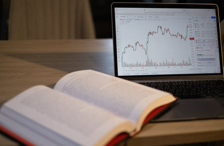 Laptop with stock market chart on screen and open book on tableの写真素材