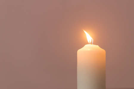 Blurred photo of single burning candle flame or light is glowing on a white candle on cream color background on table in church for Christmas, funeral or memorial service.の写真素材