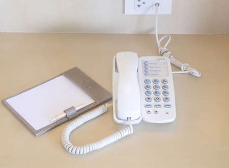 Telephone with note paper and pencil near bed is served for guests in hotel room.