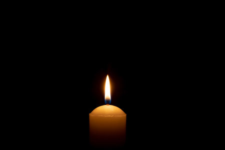 Single burning candle flame or light is glowing on a big white candle on black or dark background on table in church for Christmas, funeral or memorial service with copy space.の写真素材