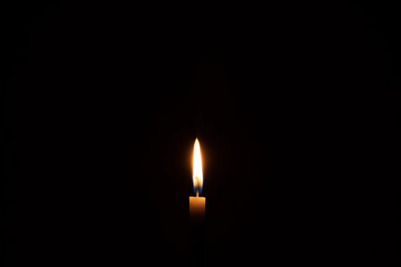 Single burning candle flame or light is glowing on a small white candle on black or dark background on table in church for Christmas, funeral or memorial service with copy space.の写真素材