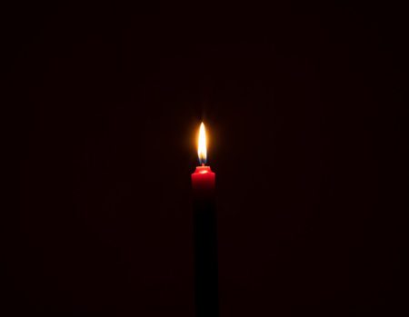 Single burning candle flame or light is glowing on a small red candle on black or dark background on table in church for Christmas, funeral or memorial service with copy space.の写真素材