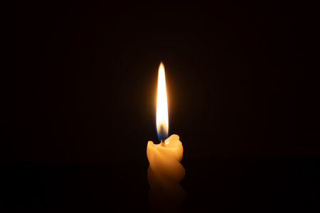 A single burning candle flame or light is glowing on a spiral white candle on black or dark background on table in church for Christmas, funeral or memorial service with copy spaceの写真素材