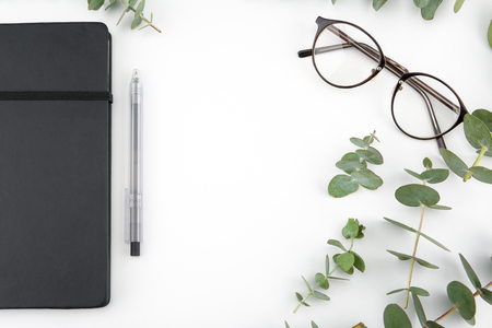 Black notebook and glasses decorated with baby eucalyptus leaves on white background with copy space in the centerの写真素材