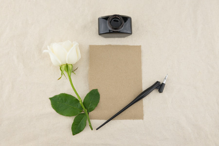 Blank brown card, oblique pen and bottle of ink decorated with white rose on white muslin fabric with copy spaceの写真素材