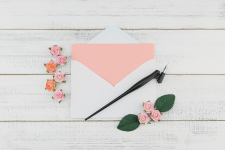 Blank pink card in white envelope and oblique pen decorate with pink rose paper flowers on white wood background with copy spaceの写真素材