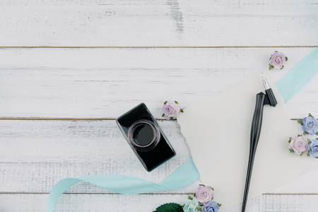 Blank white card, oblique pen and bottle of ink decorate with blue tone paper flowers and ribbon on white wood background with copy spaceの写真素材