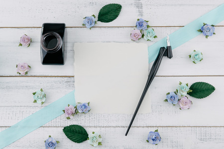 Blank white card, oblique pen and bottle of ink decorate with blue tone paper flowers and ribbon on...