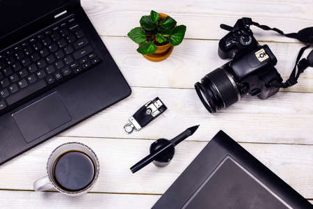 White working table with office equipment. Flat lay Laptop, digital photo apparatus, drawing tablet, pen, flash, green leaves and coffee cups on old white wooden background.の写真素材