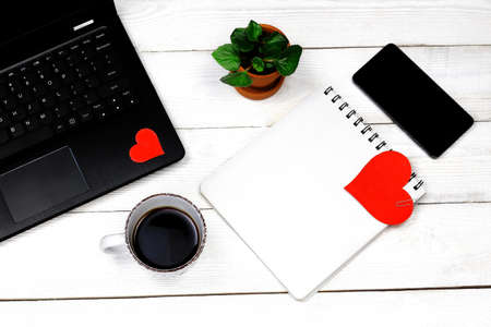 Laptop with red paper heart, notebook, pan, phone, hand clock, green leaves, womanâs hand with coffee cup on old white wooden background. Romantic Work Table concept.の写真素材