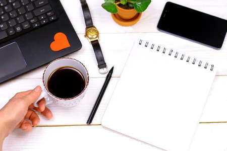 Laptop with red paper heart, notebook, pan, phone, hand clock, green leaves, womanâs hand with coffee cup on old white wooden background. Romantic Work Table concept.の写真素材