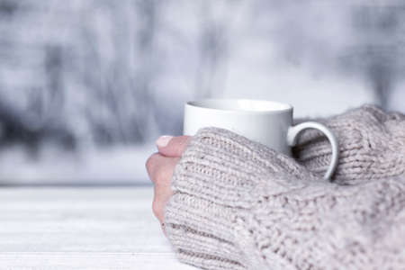 Woman's hands in sweater holding cup with hot drink and blurred winter landscape colors on background. Winter comfort concept.の写真素材