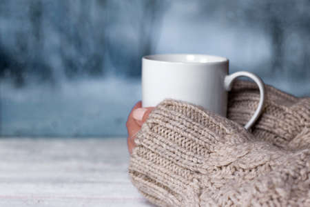 Woman's hands in sweater holding cup with hot drink and blurred winter landscape colors on background. Winter comfort concept.の写真素材