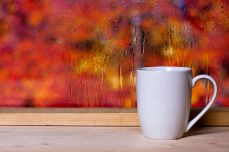 White cup on wooden background at wet window glass with blurred fall colors landscape. Autumn comfort concept.の写真素材
