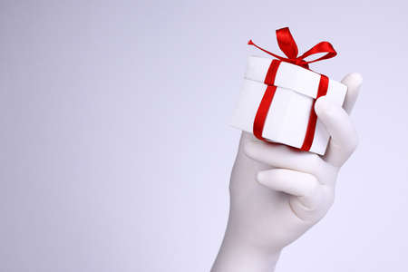 Gift box in a hand. Hand with white glove holding white gift box with red ribbon. Christmas and New Year holidays concept on white background.の写真素材