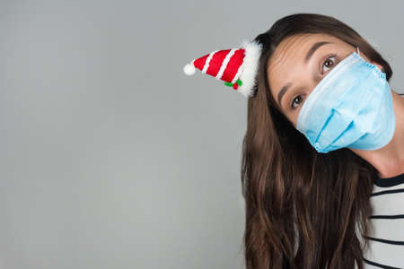 A young woman in a medical mask peeks out from behind the frame. Christmas time during the...