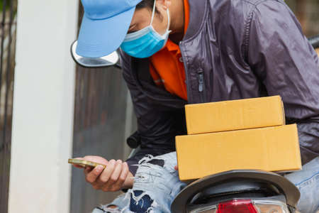 focus at cardboard box on motorcycle with background of delivery man employee wear blue cap and hygienic mask looking to mobile phone sitting on padの写真素材