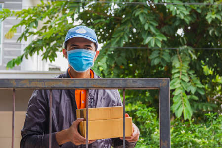 focus on cardboard box put on metal fence of house with asian delivery man wear hygienic mask and...