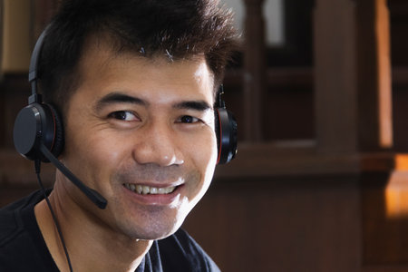 Asian man smile and wear black T-shirt use black headphone  for work from home via conference call and looking at cameraの写真素材