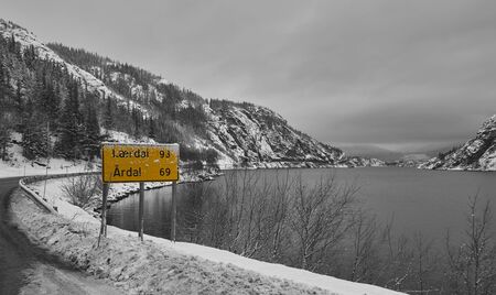 A yellow road sign showing distance to two towns on the bank of lake Tyin in Norway on a grey cloudy day. There's snow on the banks, but the lake is still free of ice.の写真素材
