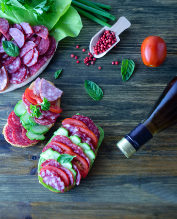 sandwich with salami with tomatoes and cucumbers next sliced salami and tomato and Basil leaves with salad and a bottle of wine on a wooden background elective focusの写真素材