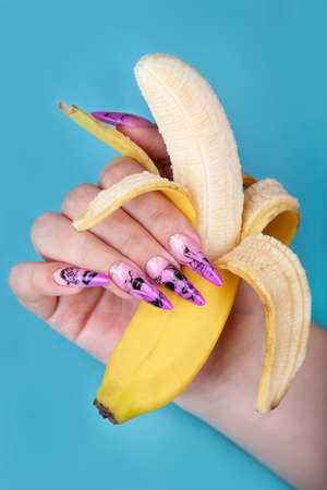 Hand with long artificial manicured nails colored with pink nail polish with summer design holding a banana. Fashion and stylish manicure.の写真素材