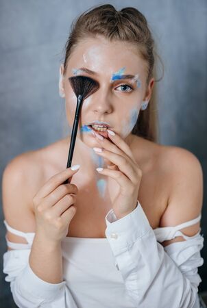Creative fashion beauty portrait of a beautiful young blond woman with a brush for cosmetics. Model girl with professional makeup and body art on a gray background.の写真素材