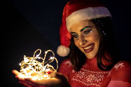 A brunette girl in a Santa Claus hat, at night on a blue background, holds lights in her hands and...