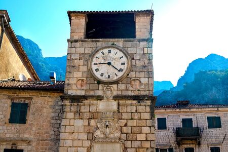 View of the clock tower in the old town of Kotor, Montenegro. We see old stone houses with red roofs, a clock tower, in the background a mountainの写真素材