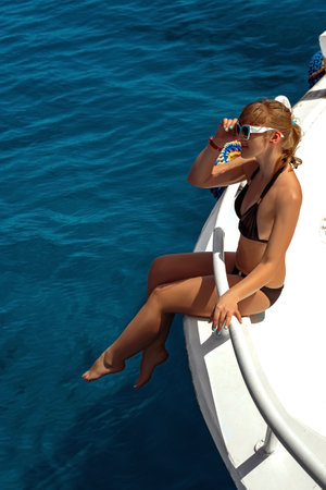 A beautiful blonde woman in a black bikini and white shirt is enjoying her summer vacation sunbathing on a luxury yachtの写真素材