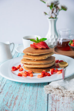 Homemade pancakes with strawberries and honey on a white wooden background.