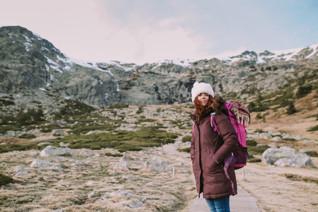 Brunette girl with a hat on her head and a travel backpack contemplates the landscape that surrounds her from a valley next to the mountainsの写真素材