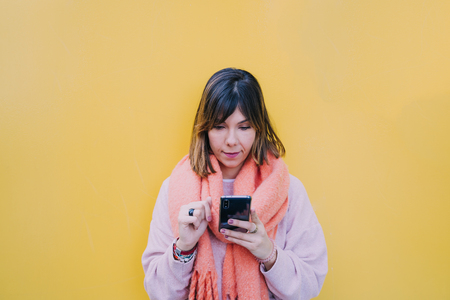 Woman with short brown hair holds the phone with her hands. Look at the phone carefully while typing with your fingers. He is surfing the internet quietly. It rests on a yellow wall.の写真素材