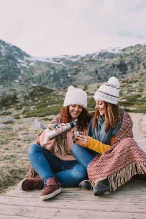 Two friends wrapped in a blanket are sitting in the meadow while they take out a cup to prepare a brothの写真素材