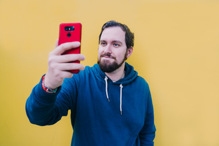 Dark-haired man with blue eyes looks at his cell phone while taking a picture. Hold the phone with one hand and hold the arm upward. He has a beard, a blue sweater and the phone is redの写真素材