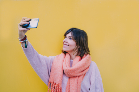 Brunette woman is in the street next to a yellow wall and a picture is taken with her mobile. Smile at the camera while holding the phone with your hand extended. He has short hair and wears a coral colored scarf.She smiles at the cell phone screen.の写真素材