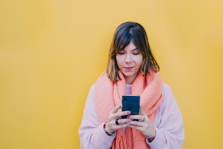 Woman with short brown hair holds the phone with her hands. Look at the phone carefully while typing with your fingers. He is surfing the internet quietly. She smiles at the cell phone screen.It rests on a yellow wall.の写真素材