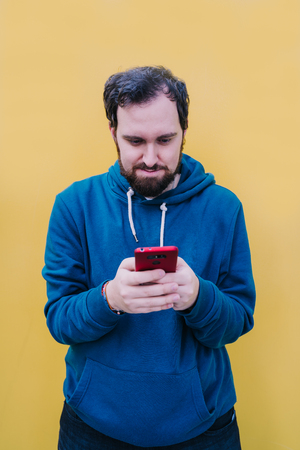 Young boy is in the street next to a yellow wall and holds his mobile with both hands. Look carefully at the screen of the mobile watching something. He has dark hair and a beard. He wears a blue sweater and the phone is red.の写真素材