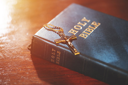 A bronze crucifix necklace resting on a closed Holy Bible, illuminated by warm light, symbolizing Christian faith, the teachings of Jesus, and spiritual devotion.の写真素材