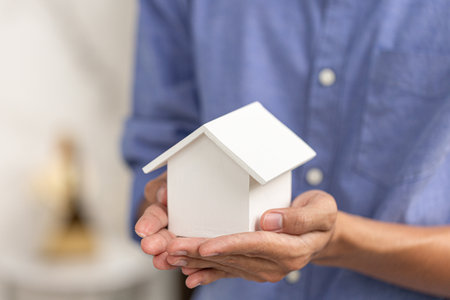 Person holding a small white wooden house with both hands, symbolizing home ownership, real estate, mortgage, investment, and property insurance.の写真素材