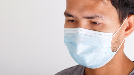 A close-up of an Asian man wearing a light blue surgical mask, showing a serious expression. The image emphasizes public health, safety, and personal protection during illness or pandemic situations.の写真素材