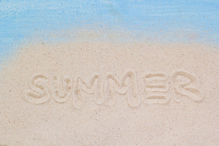 The word "SUMMER" is written in soft beach sand with a light blue wooden background, evoking a bright and cheerful tropical vibe.の写真素材