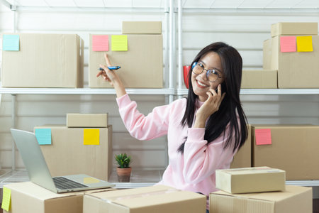 Smiling young Asian woman running small online business at home, using laptop and phone while managing orders. Surrounded by cardboard boxes, colorful sticky notes, and plants, in a modern workspace environment.の写真素材