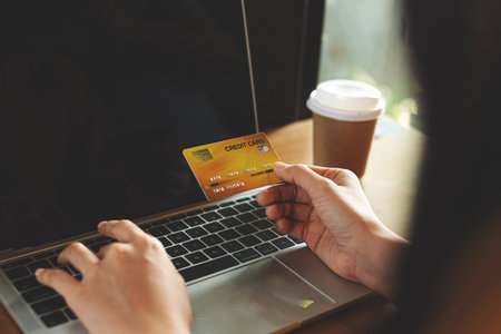 Close-up of a woman holding a credit card while using a laptop on a wooden table with a coffee cup beside. Concept of online shopping, digital payment, e-commerce, and secure transactions.の写真素材