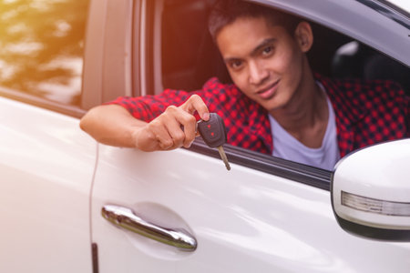 Young man sitting in a car, smiling and holding a car key out of the window, symbolizing car ownership, freedom, or a new purchase.の写真素材