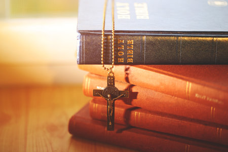 A crucifix necklace hanging on stacked Holy Bibles, illuminated by soft sunlight, symbolizing...