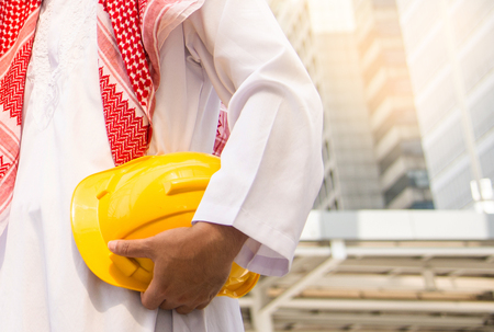 Arab Engineer or Safety officer holding helmet construction or yellow helmet for workers security with city background in construction site.の写真素材