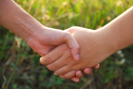 Hands of two young women expressed great friendship together by holding hands in the evening sunshine on blurred grass background.の写真素材