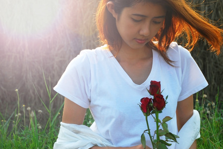 Asian young woman are moment in love. She sit on the field in the evening sunshine have a mild breeze and holding a red rose in hand.の写真素材